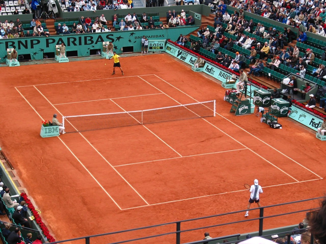 法國網球公開賽（Roland-Garros）紅土場地比賽實景，球員於中央球場對戰，觀眾席座無虛席，場邊可見 IBM、Lacoste 與 Perrier 等賽事贊助品牌，充分展現紅土網球特有的比賽氛圍與文化象徵。